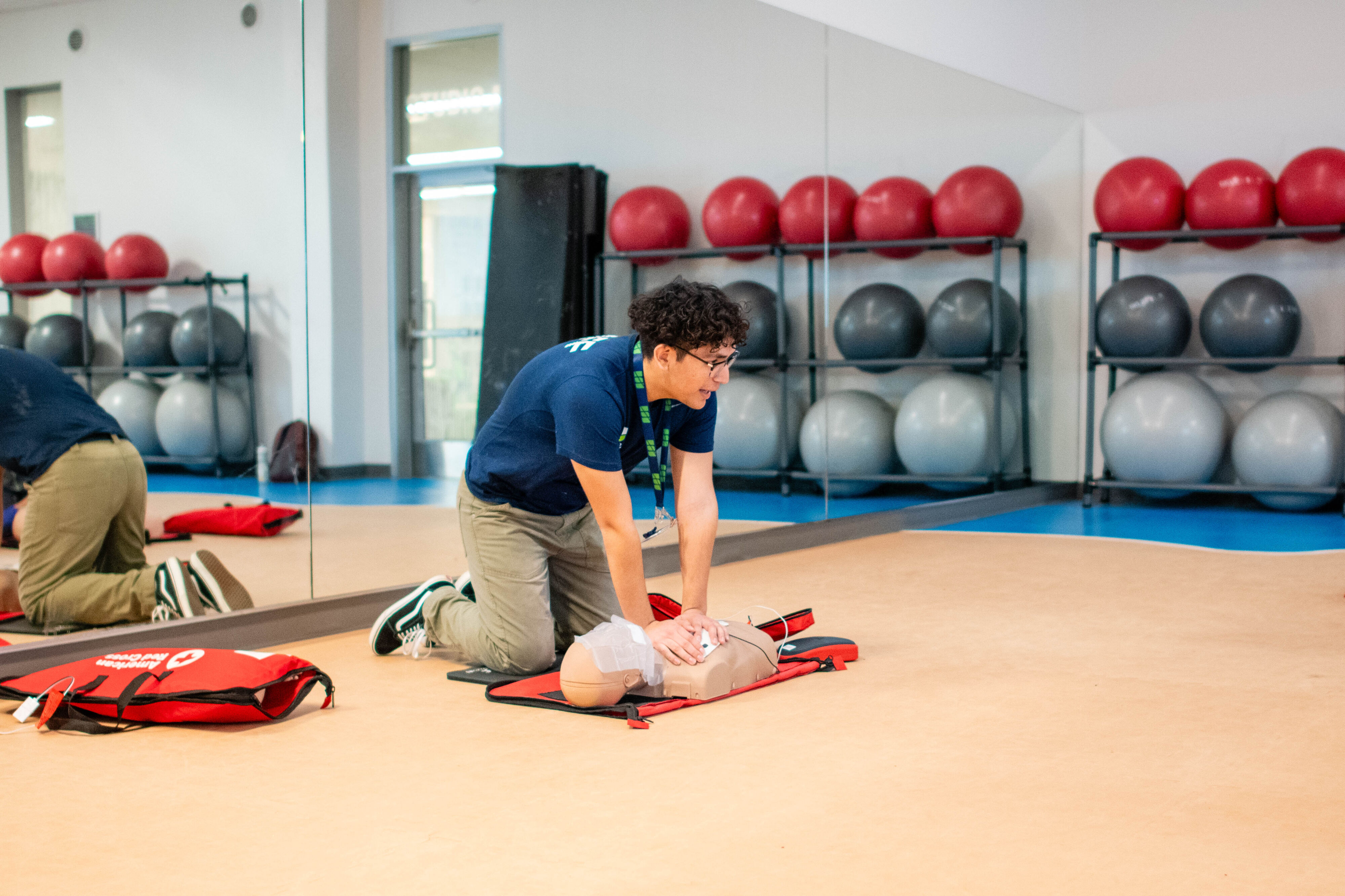  An individual is kneeling to perform CPR on a training dummy in a gym