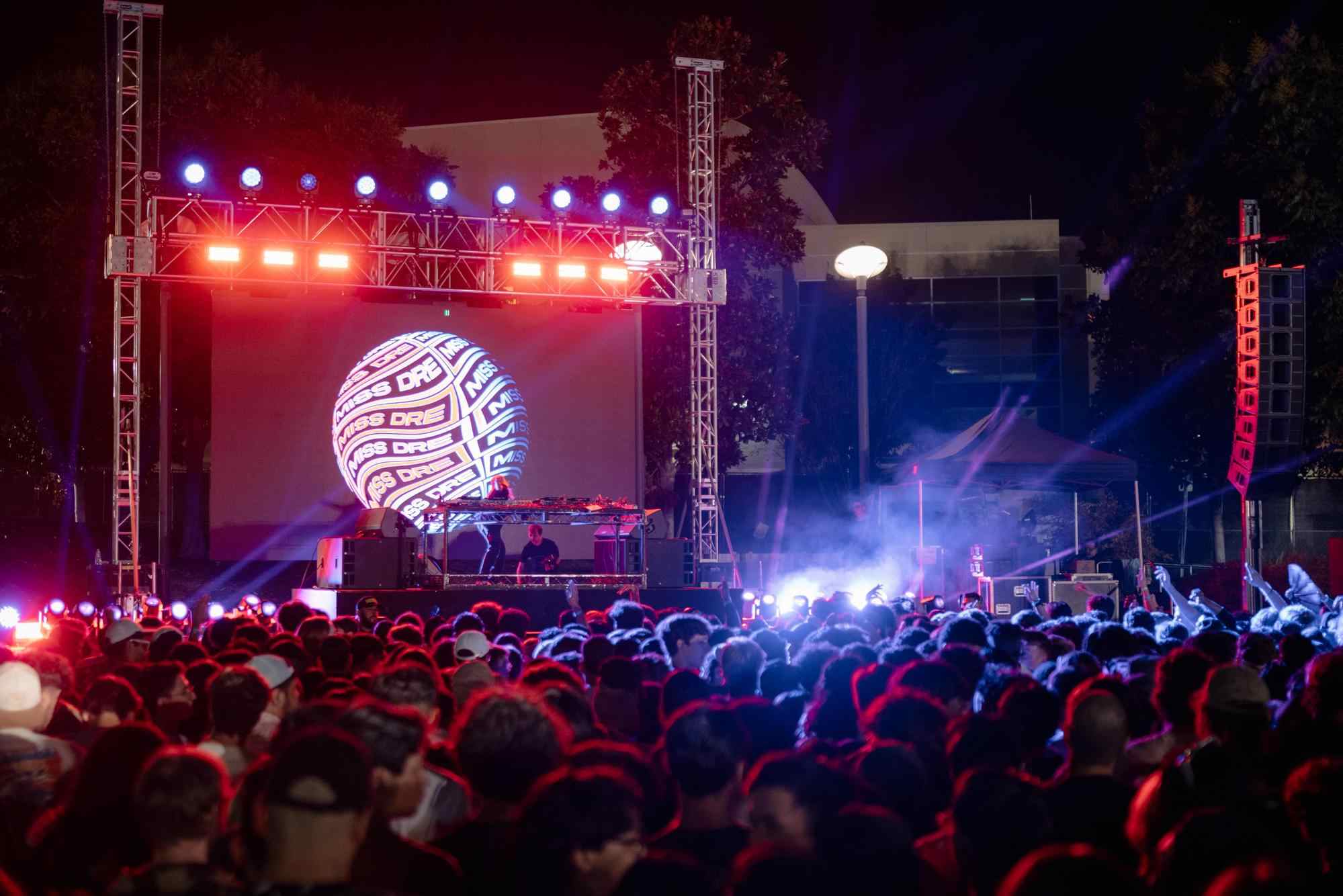 large crowd at an outdoor music event at night with a DJ performing on stage