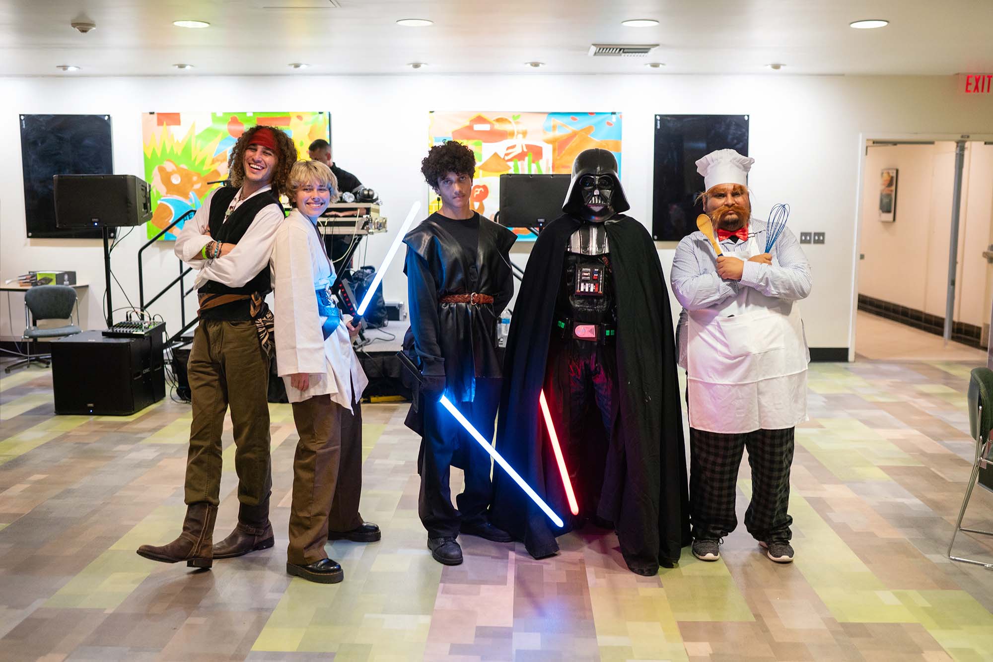  A group of people in various costumes are standing indoors, with two holding lightsabers, one dressed as a chef, and one in a dark cape and mask. There's a music setup in the background.