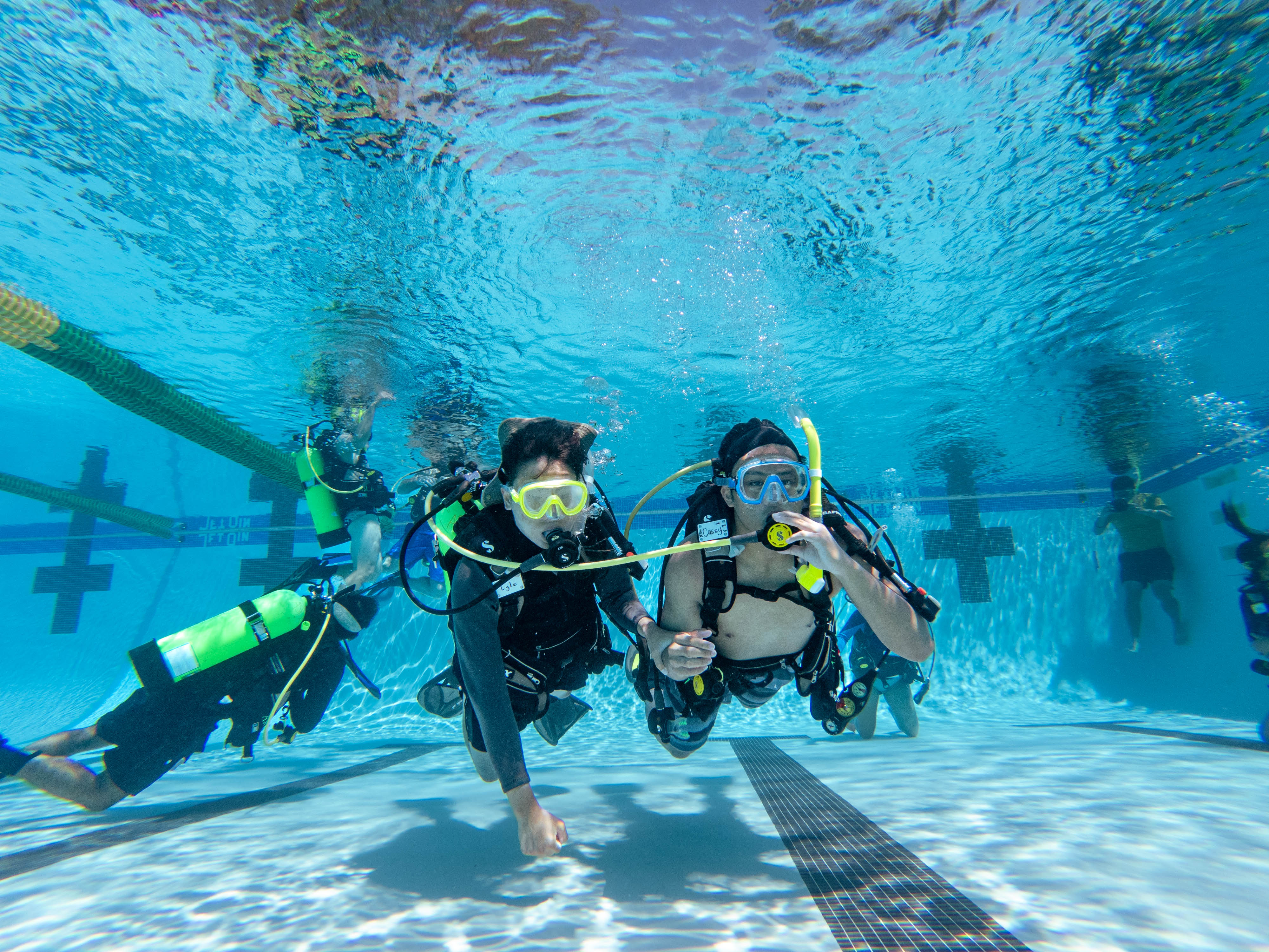Two scuba divers with masks and snorkels are underwater in a clear pool, with other divers and swimming pool lanes visible in the background.