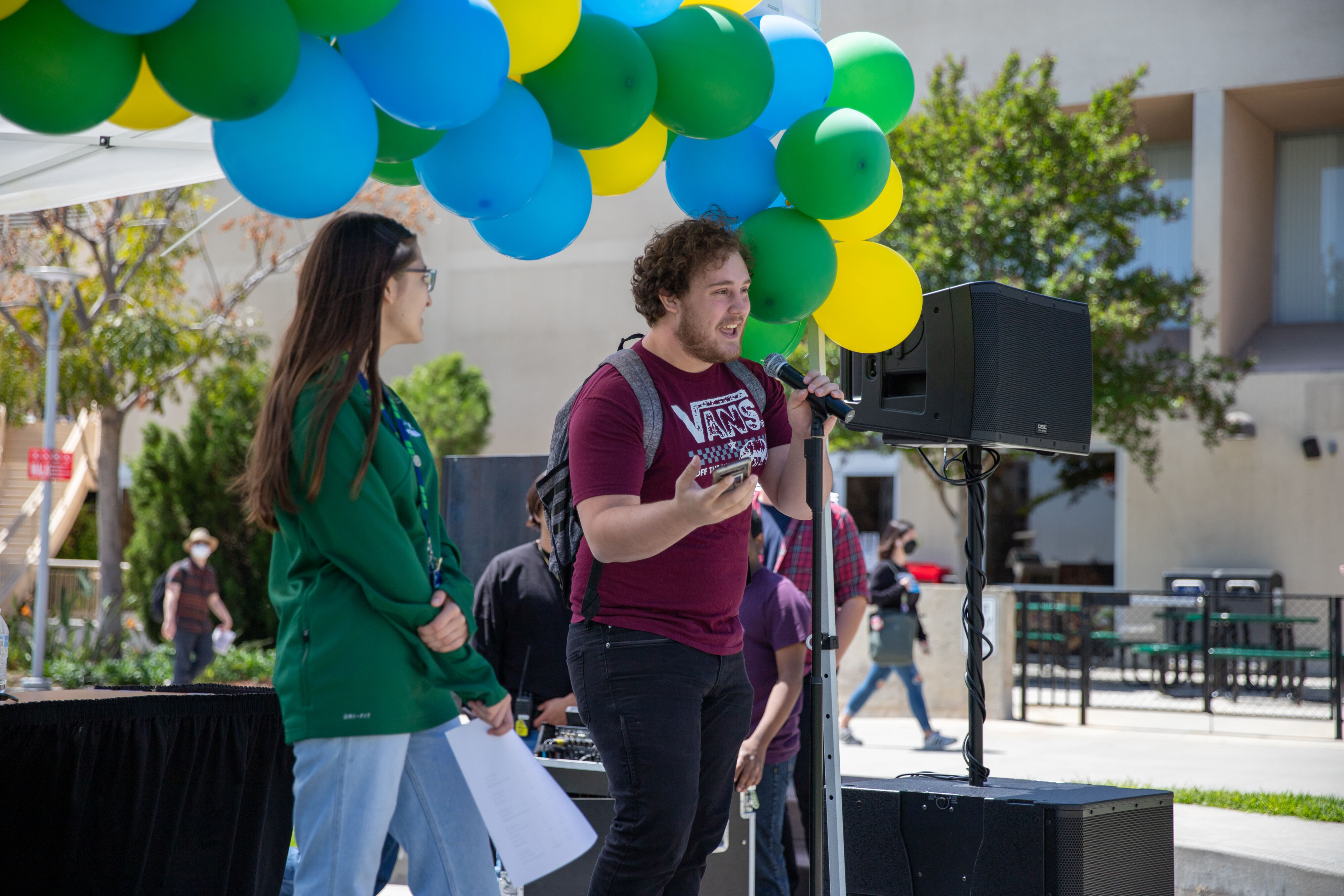 Alt: A student speaking into a microphone on a stage with yellow, green, and blue balloons 