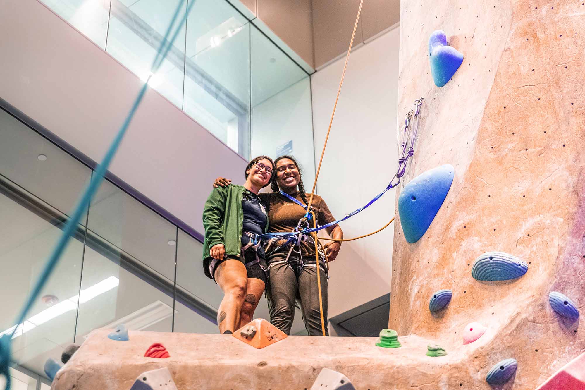 Two climbers are smiling and posing at the top of an indoor climbing wall
